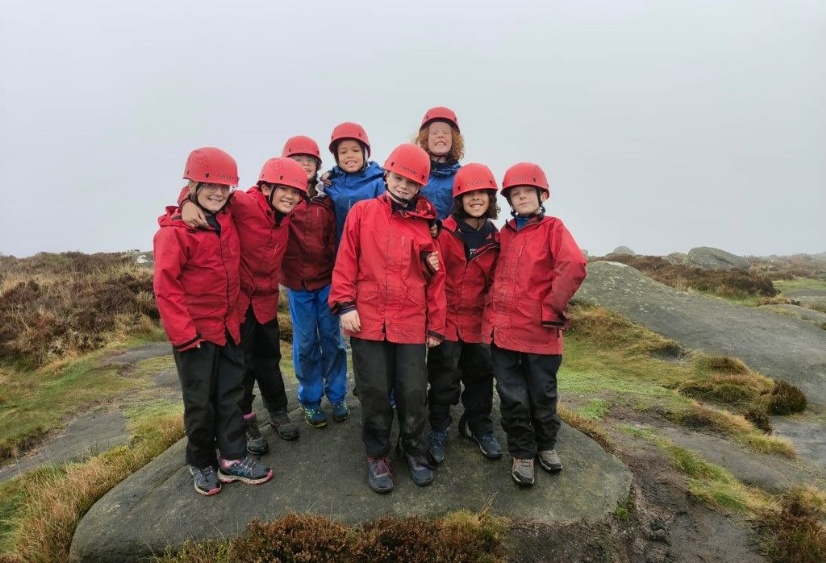 A group of boys and girls in hard hats and overalls stand at the top of a rocky outcrop in the Peak District